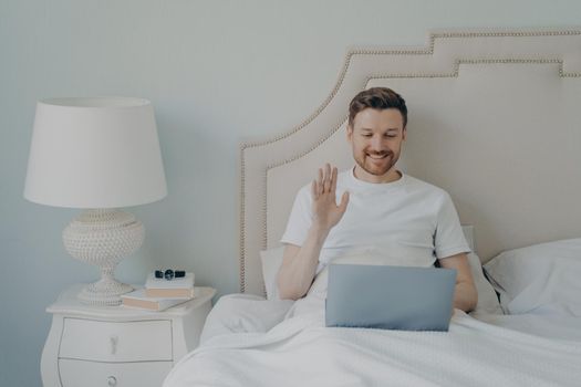 Happy young man with stubble waving hand to laptop while having online video call and sitting in bed, making conversation with his close friends during leisure time on weekend at home