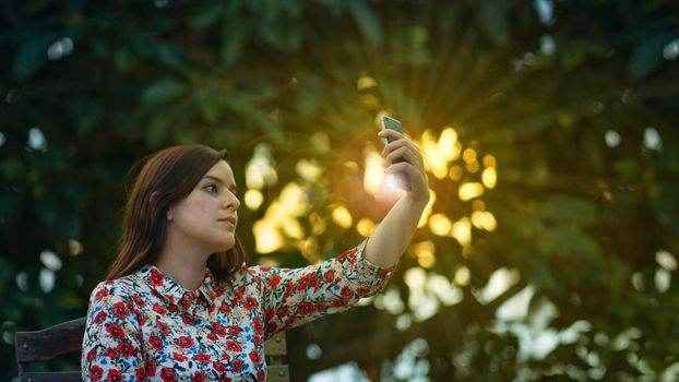 Beautiful young latin woman in floral design dress taking a selfie with her smartphone in the garden at sunset with the sun's rays passing through the trees in the background