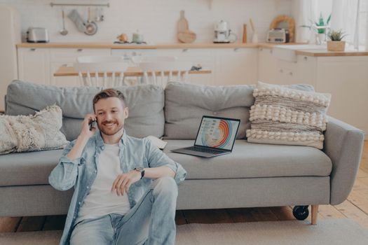 Confident male freelance worker calling his employer to tell him good news about project, making another client satisfied, laptop with infographics, sitting on floor while resting against couch