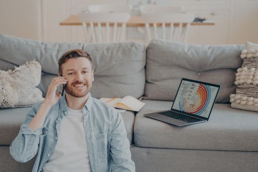 Young smiling male freelance worker calling his client to notify him about finished project, happy to receive payment, sitting on carpet while resting against couch, blurred background
