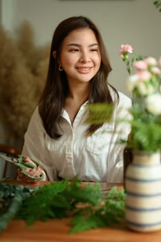 Portrait of smiling female florist making bouquet for a client at wooden counter. Floristry, small business and people concept.