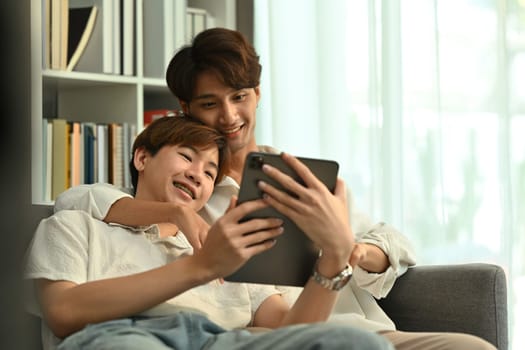 Passionate young gay couple relaxing on couch and browsing internet on digital tablet. LGBT, love and lifestyle concept.