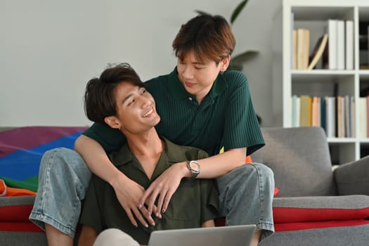 Romantic happy gay couple embracing and using laptop in living room. LGBT, love and lifestyle relationship concept.