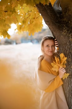 The blonde stands near the autumn tree. Thoughtful woman looks ahead, dressed in a yellow dress. Autumn content.