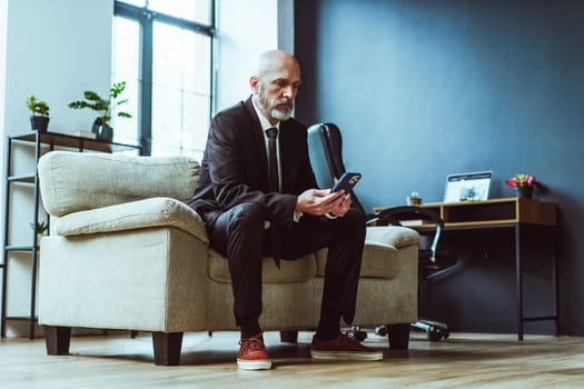 Old businessman dressed in black suit can be seen either texting on phone or browsing internet. Mature man, with distinguished silver beard, sits comfortably in chair within stylish loft office. . High quality photo