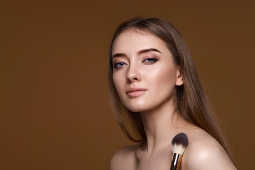 young woman applying blush on cheeks with make-up brush on beige background. copy space