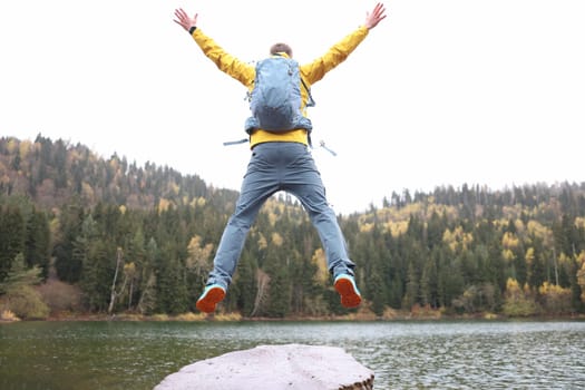 Happy male traveler with backpack jumping near forest lake back view. Travel and goal achievement concept