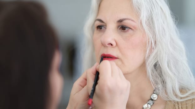Cosmetologist painting lips with red lipstick to elderly woman. Makeup for any age concept