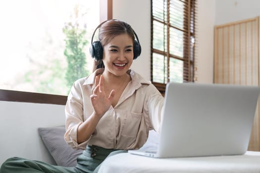 Young happy smiling woman in casual clothes sitting on sofa work online talk by video call with laptop pc computer wave hand rest relax indoors at home People lifestyle leisure concept.