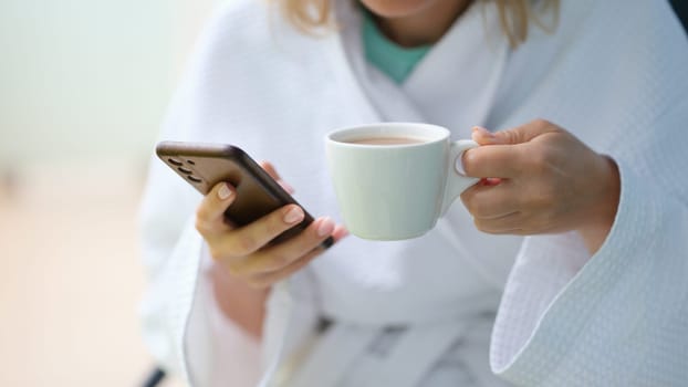 Woman in white bathrobe drinking coffee from cup and holding mobile phone at home closeup. Blogging social media concept