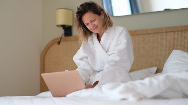 Young woman in white bathrobe working remotely with laptop in bed at home. Telework concept