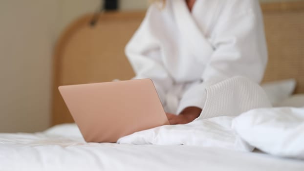 Woman in white bathrobe sitting with laptop in bed at home closeup. Remote work online concept