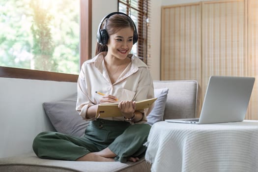 woman taking notes in notebook while using laptop at home in living room. Focused lady writing in notepad.