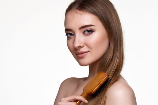 Beautiful smiling brunette woman looks at the camera and combs her hair with wooden brush isolated on white background