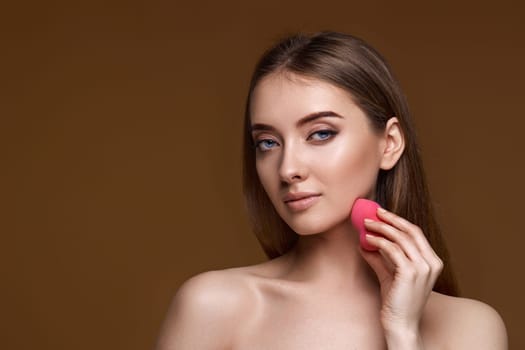 smiling young woman applying foundation on skin with beauty blender isolated on beige background