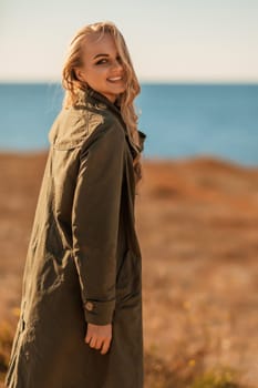 Portrait blonde sea cape. A calm young blonde in an unbuttoned khaki raincoat stands on the seashore, under the raincoat there is a black skirt and top.