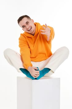 young man in orange sweatshirt sitting on white cube on white background. Full length