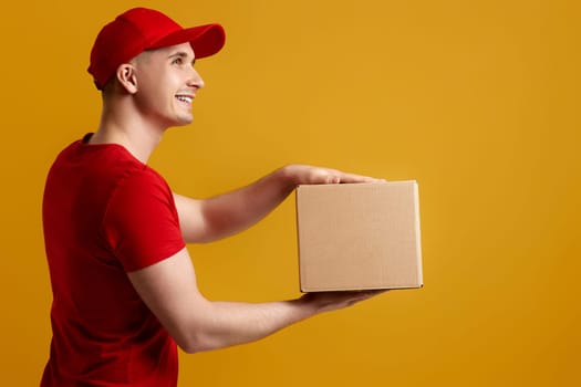 Delivery man employee in red cap, red t-shirt holding cardboard box isolated on yellow background