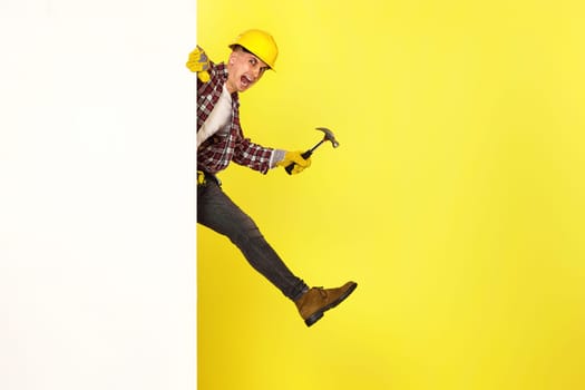 professional builder in work clothes, helmet holds hammer and comes out from behind the banner. copy space for text