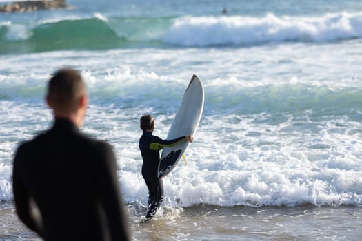 Smiling son on the shoulders of his father walking in the sea wearing wetsuits. Mid shot