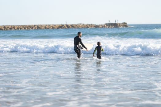 A family of surfers - father and son in wetsuits go to sea. Mid shot