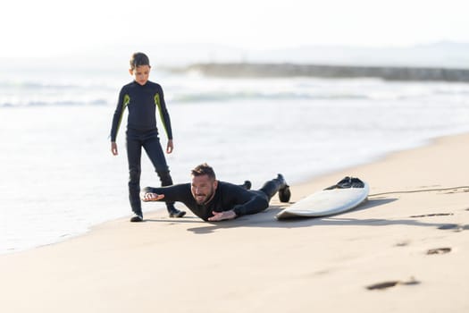 A family of surfers - smiling father lying on the sand by the surfboard showing his son how to lie on the board. Mid shot