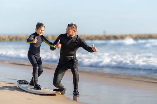 A little boy stands on the surfboard on the seashore and his father standing by him - showing thumbs up. Mid shot