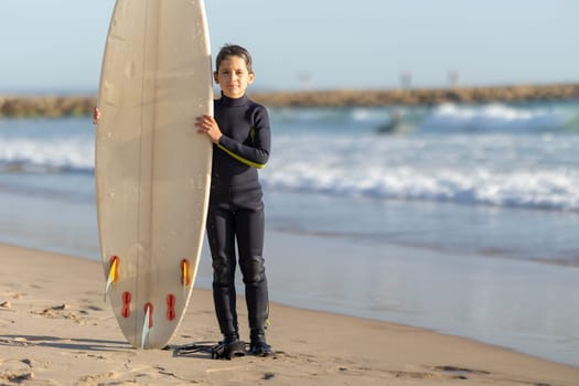 A little boy standing on the seashore with a big surfing board. Mid shot