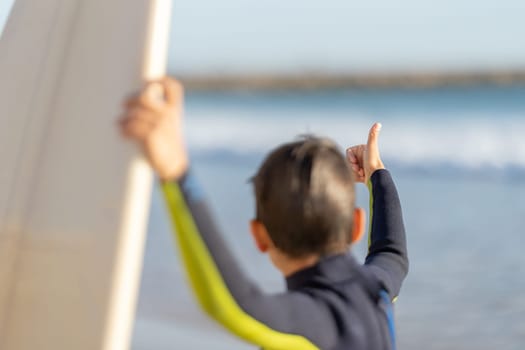 Little boy in wetsuit shows thumbs up looking at surfboard - back view. Mid shot