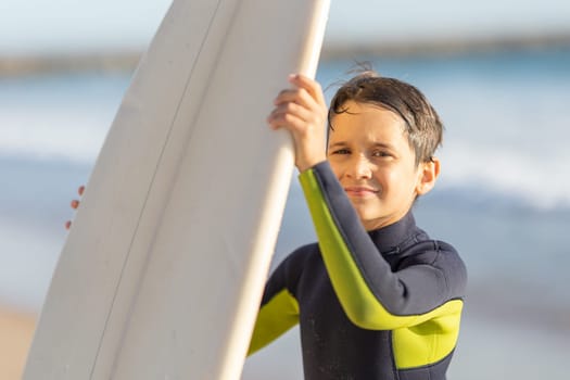 Cute little boy in a wetsuit holding a surfboard. Mid shot