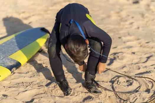 A man surfer fastens a board to his leg while sitting on the shore. Mid shot