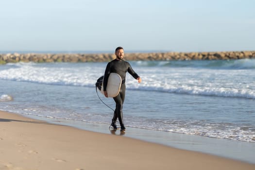 A man surfer in a wetsuit walking on the shore holding a surfboard. Mid shot