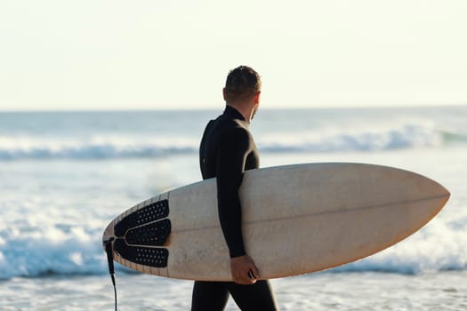 A surfer in a wetsuit walking on the shore holding a surfboard and looking at the sea. Mid shot