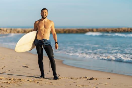 A man surfer with naked torso standing on the seashore holding a surfing board. Mid shot
