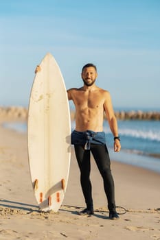 Smiling man surfer with naked torso standing on the seashore holding a surfing board. Vertical shot
