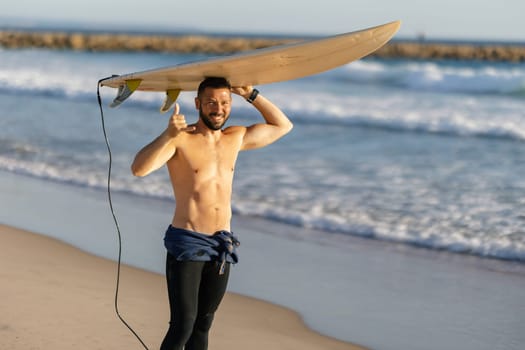A smiling man surfer with naked torso holding a surfing board over his head and showing a thumb up. Mid shot
