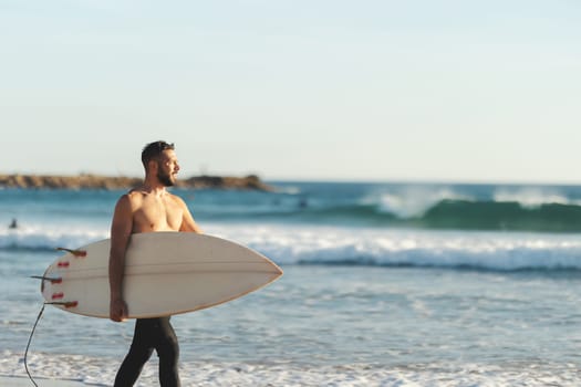 A smiling athletic man surfer with naked torso walking on the seashore. Mid shot