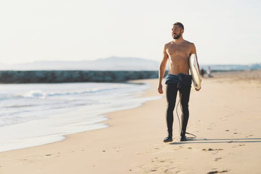 An athletic man surfer with naked torso standing on the seashore. Mid shot