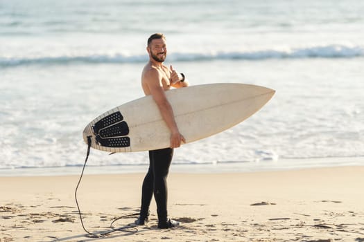 An athletic man surfer with naked torso standing by the sea and showing a thumb up looking in the camera. Mid shot