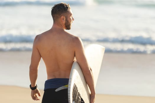 An athletic man surfer with naked torso standing by the ocean. Mid shot