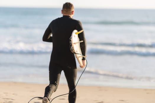 An athletic man surfer in a wetsuit running to the ocean. Mid shot