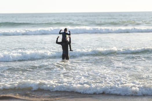 Little son on the shoulders of his father in the sea showing muscles - back biew. Mid shot