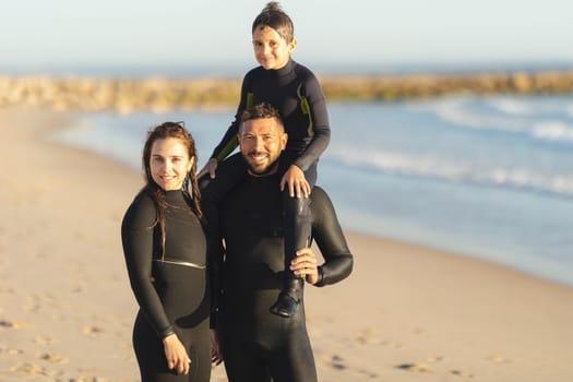 A family of surfers stands on the seashore in wetsuits. Mid shot