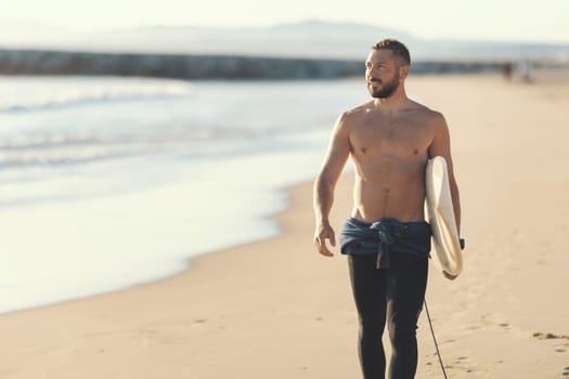 An athletic man surfer with naked torso walking on the seashore. Mid shot