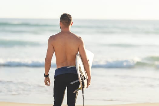 An athletic man surfer with naked torso standing by the sea - back view. Mid shot