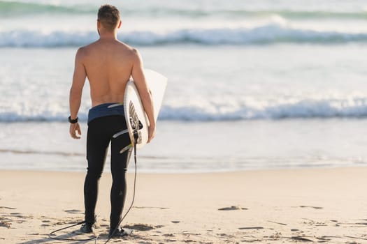 An athletic man surfer with naked torso standing by the ocean - back view. Mid shot
