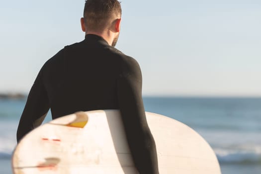 An athletic man surfer in a wetsuit standing by the ocean. Mid shot