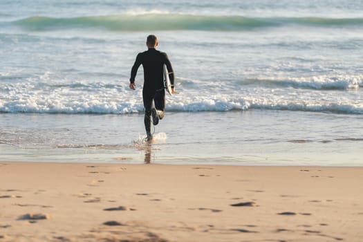 An athletic man surfer in a wetsuit running to the ocean holding a surfing board. Mid shot