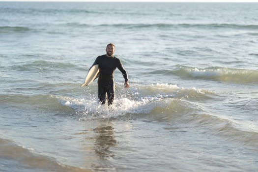 Smiling man surfer coming out of the sea holding a surfboard. Mid shot