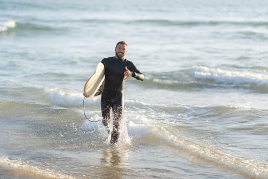 Smiling man surfer running out of the sea holding a surfboard and showing shaka. Mid shot